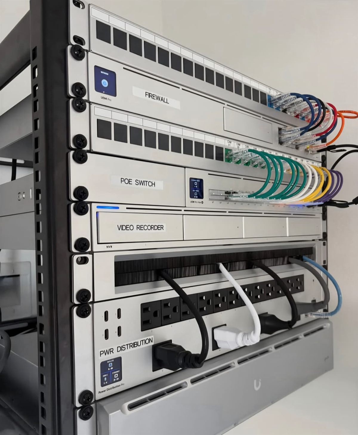 Wall-mounted Ubiquiti UniFi rack installed by Anfield IT — labeled firewall, PoE switch, video recorder, power distribution and battery backup with color-coded patching